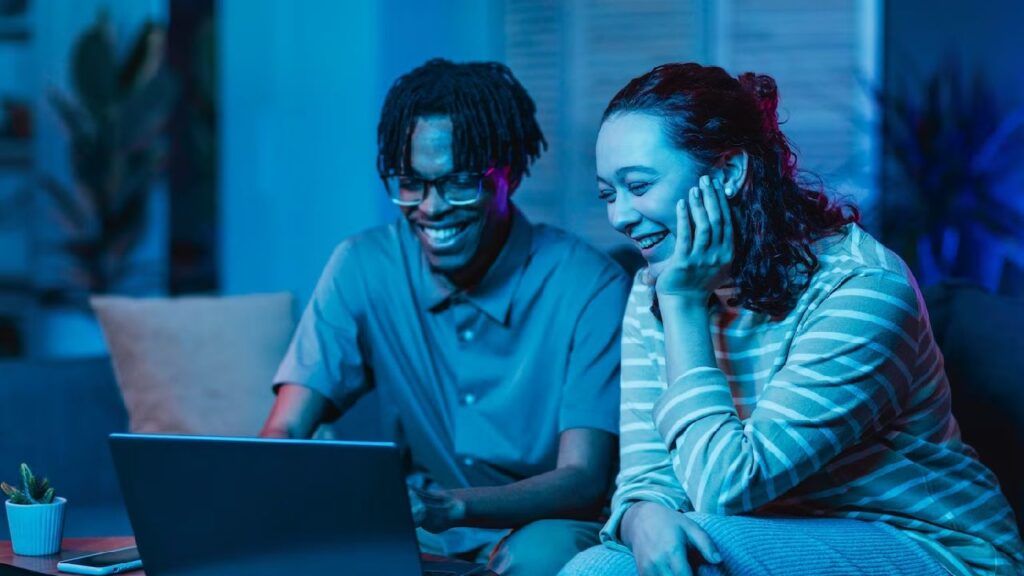 Casal sorrindo enquanto usa um laptop em um ambiente moderno e iluminado com luz azul. Eles parecem felizes e descontraídos