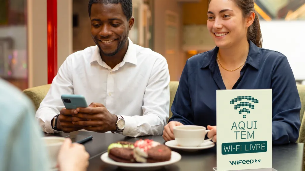 Casal de clientes em cafeteria conversando e sorrindo, enquanto uma placa indica “AQUI TEM WI-FI LIVRE” WiFeed na mesa com xícara de café