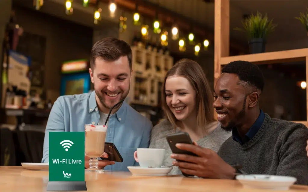 Três amigos sorrindo em um café, interagindo com seus smartphones enquanto desfrutam de bebidas. O sinal de Wi-Fi livre está visível na mesa.