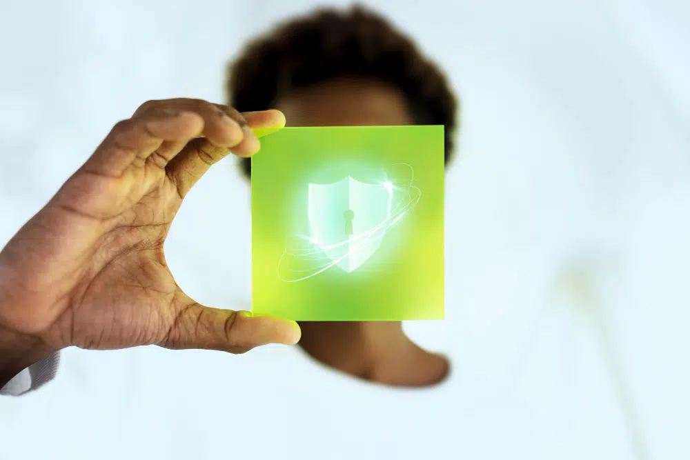 	
Pessoa segurando um cartão verde com símbolo de escudo de proteção digital, representando segurança de dados e proteção de informações online.